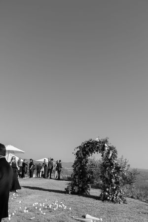 Outdoor wedding ceremony setup with floral arch against clear sky in black and white.