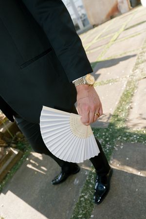 A person in formal black attire holds a white folding fan while standing on a paved surface.