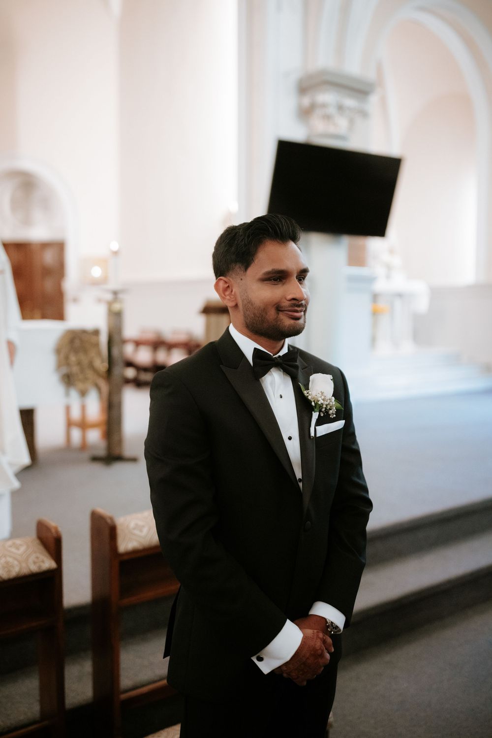 Groom in black tuxedo reacts emotionally during wedding ceremony in bright church setting.
