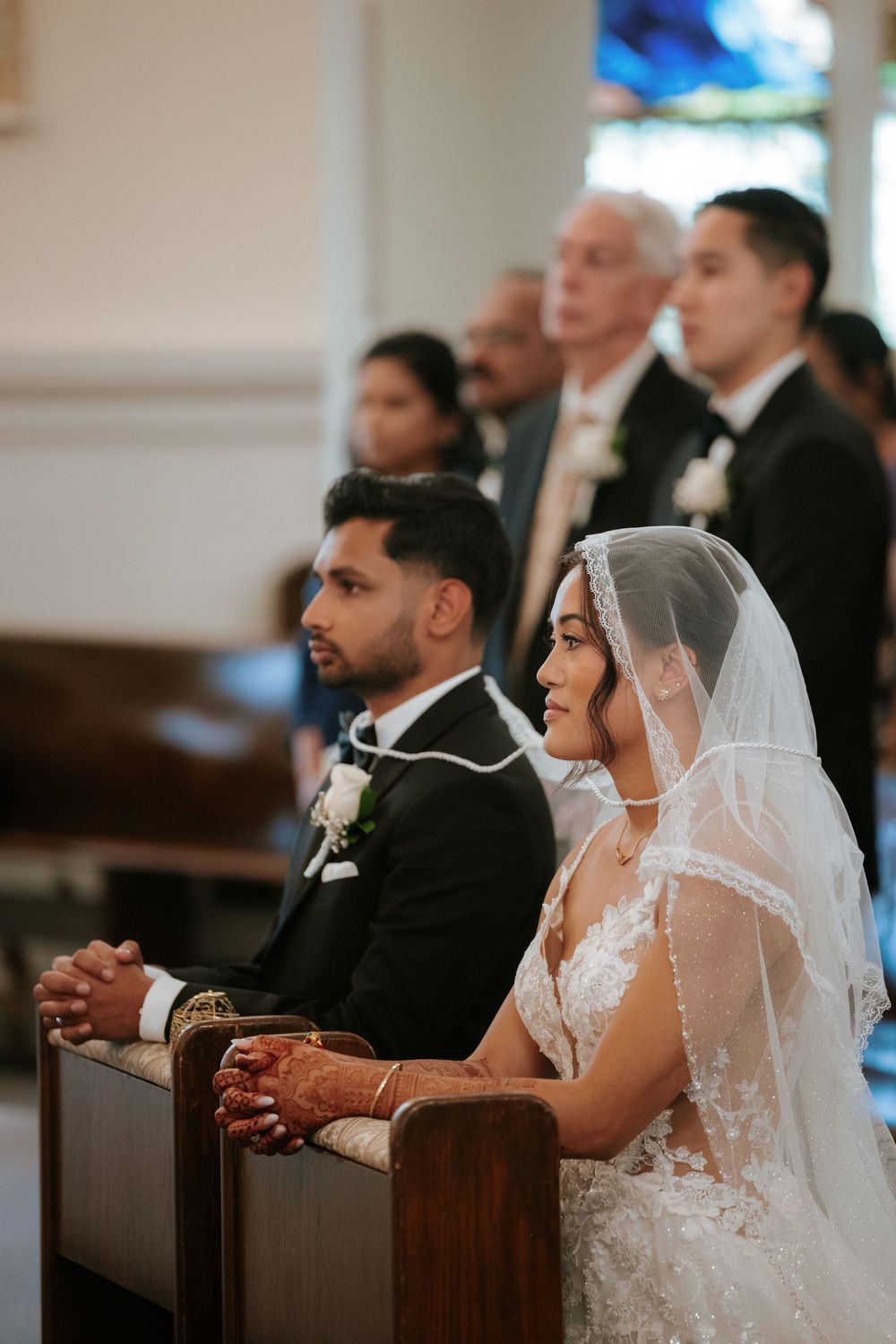 A bride in a white lace dress and veil sits in a wooden church pew during a wedding ceremony.
