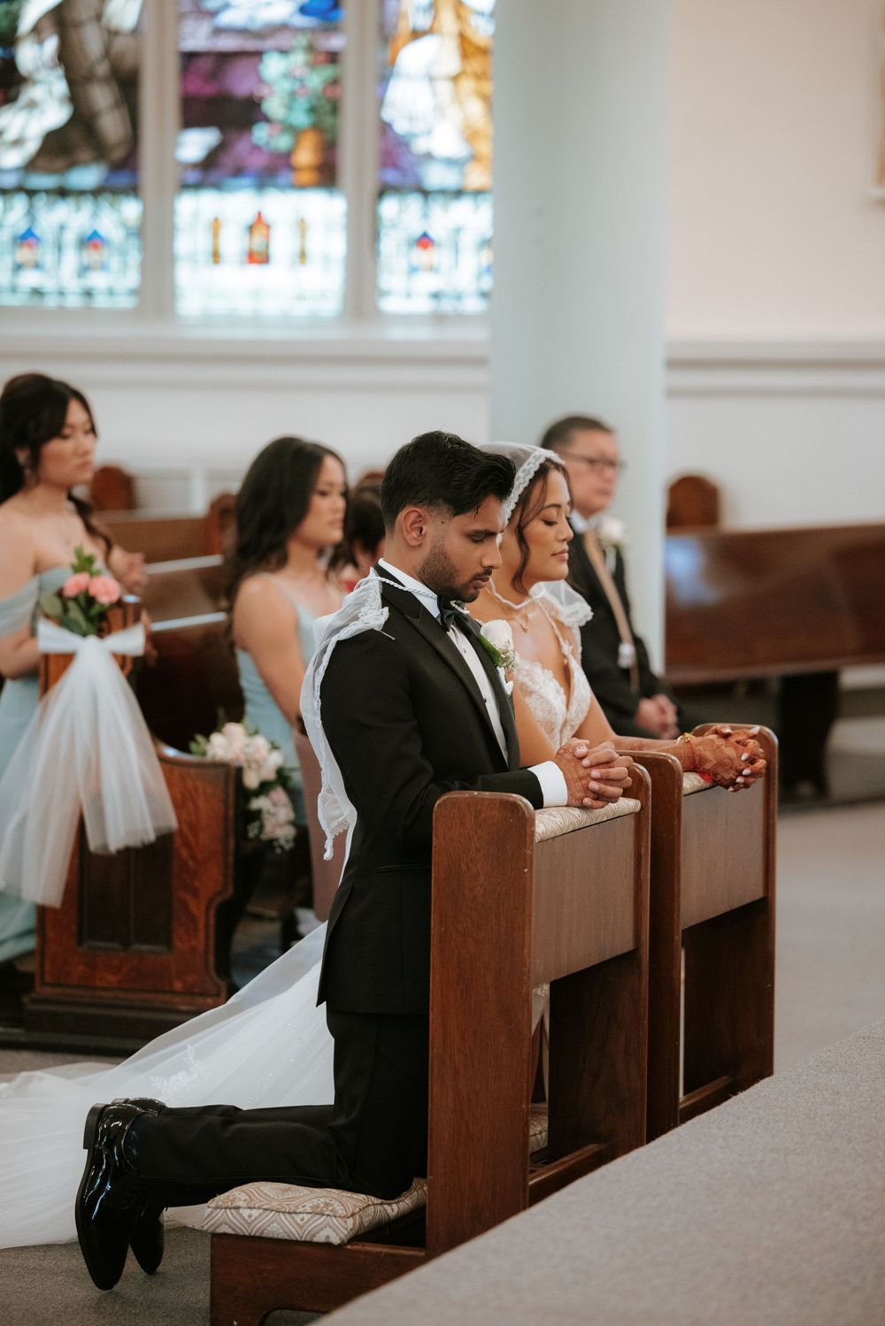 Wedding guests seated in wooden church pews during a ceremony with stained glass windows visible in background.