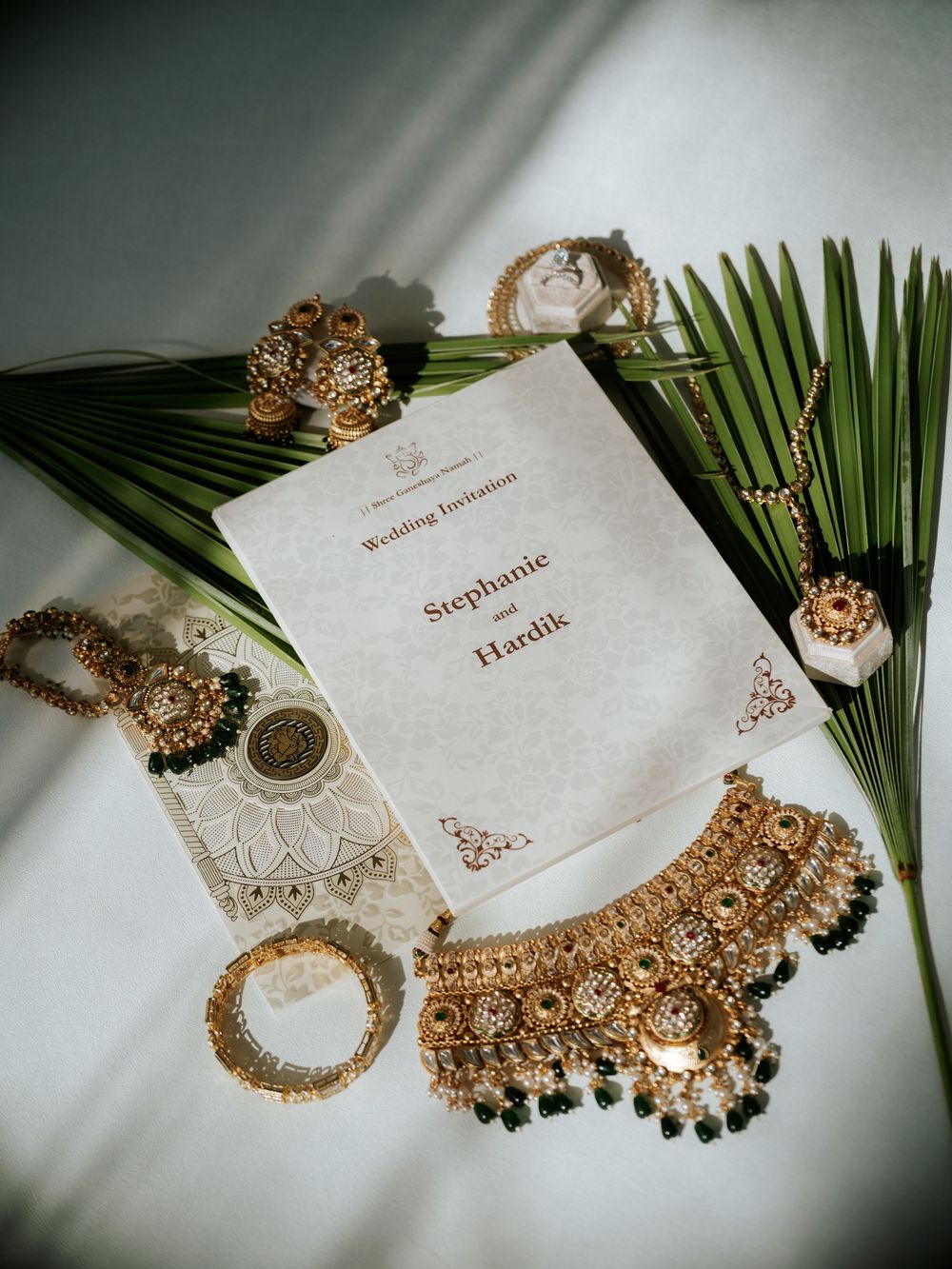 Elegant gold bridal jewelry and wedding invitation cards displayed with tropical palm leaves on white fabric background.