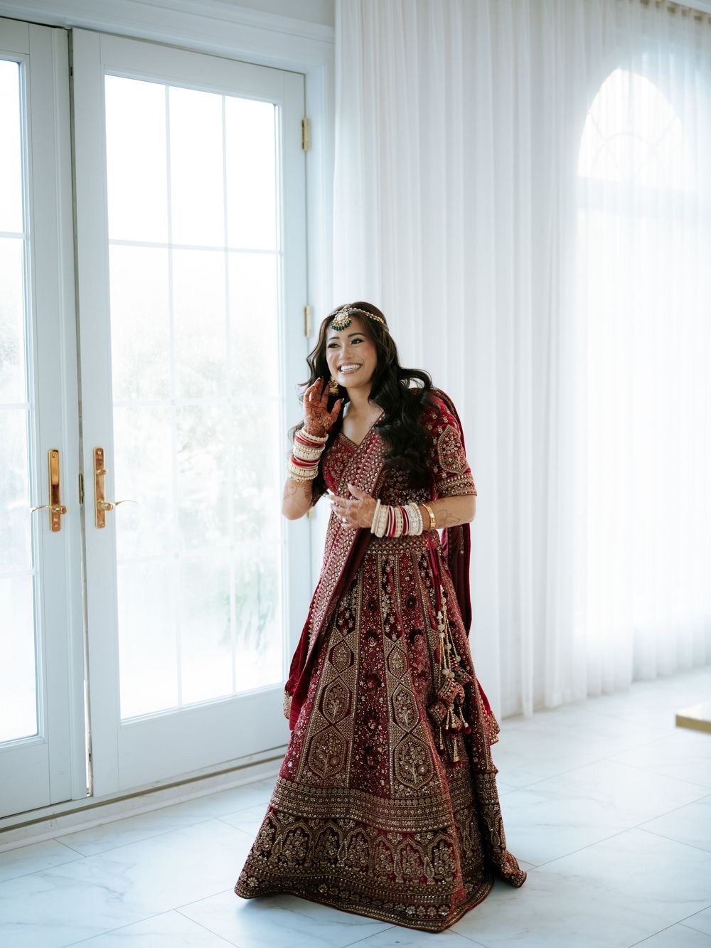A person in an ornate burgundy and gold lehenga poses gracefully in a bright white room with sheer curtains. at the Rockleigh, NJ