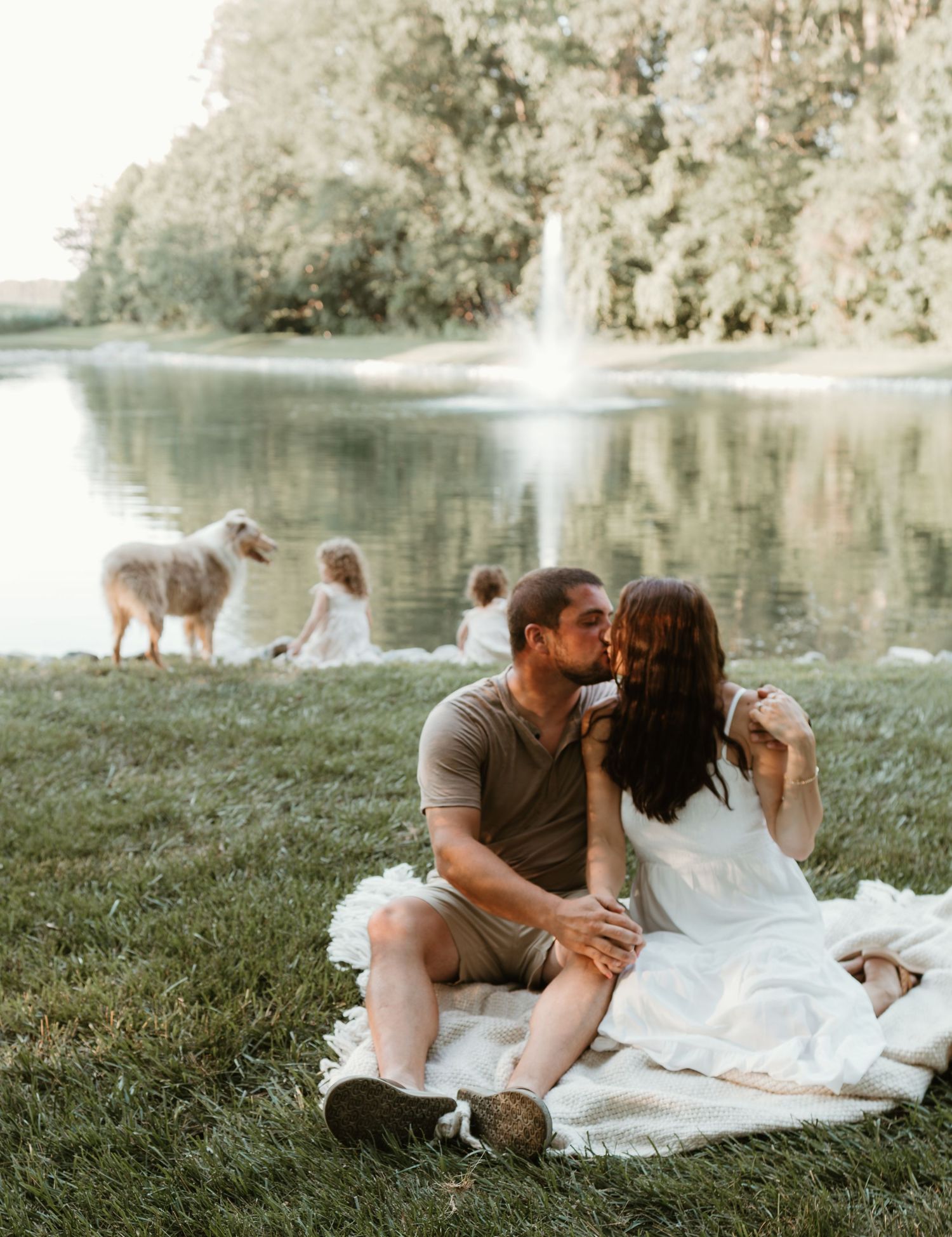 Couple enjoys romantic picnic by lake while children play in background.