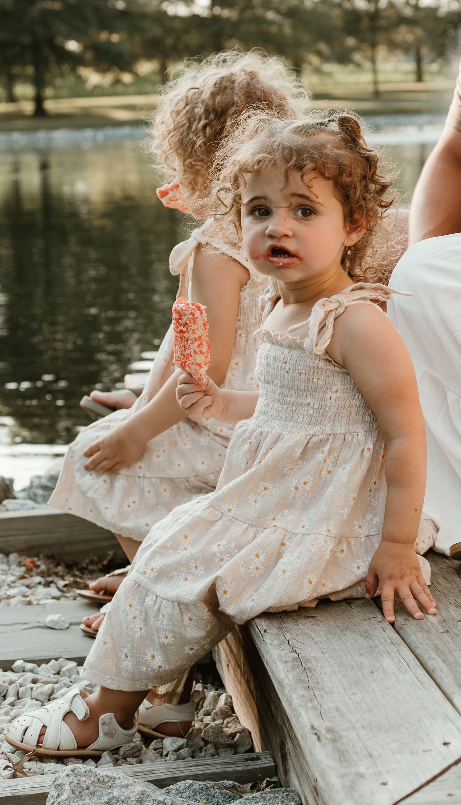 A curly-haired toddler in a white summer dress sits by a lake holding a snack.