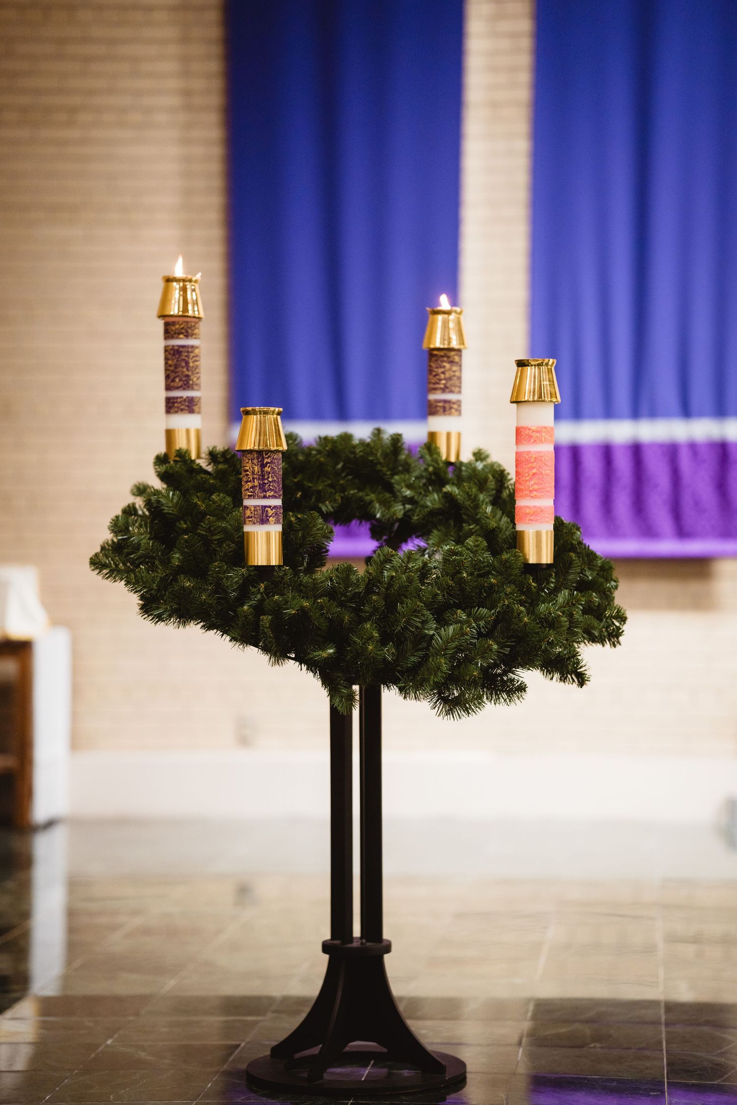 Advent wreath with golden candle holders on black stand against blue and white church backdrop.