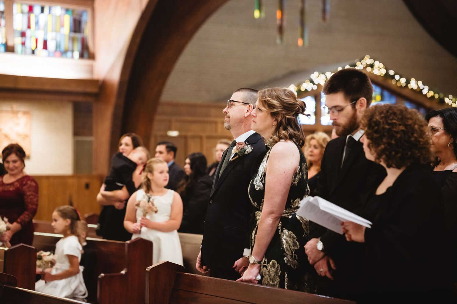 A group of people gather in a church pew during a wedding ceremony with warm lighting and festive decorations.