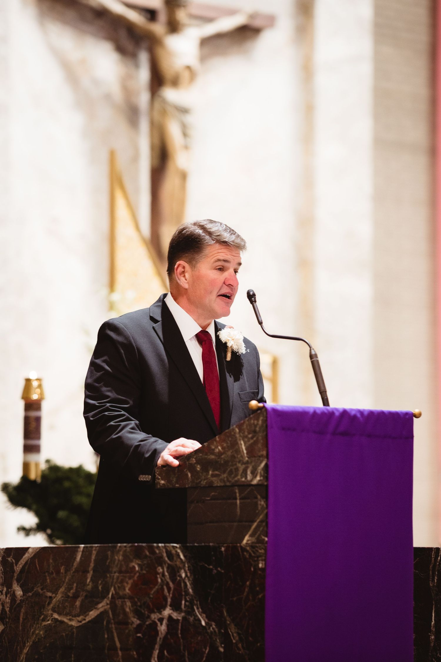 Speaker at a podium with marble backdrop delivers remarks in a dark suit and red tie during formal event.