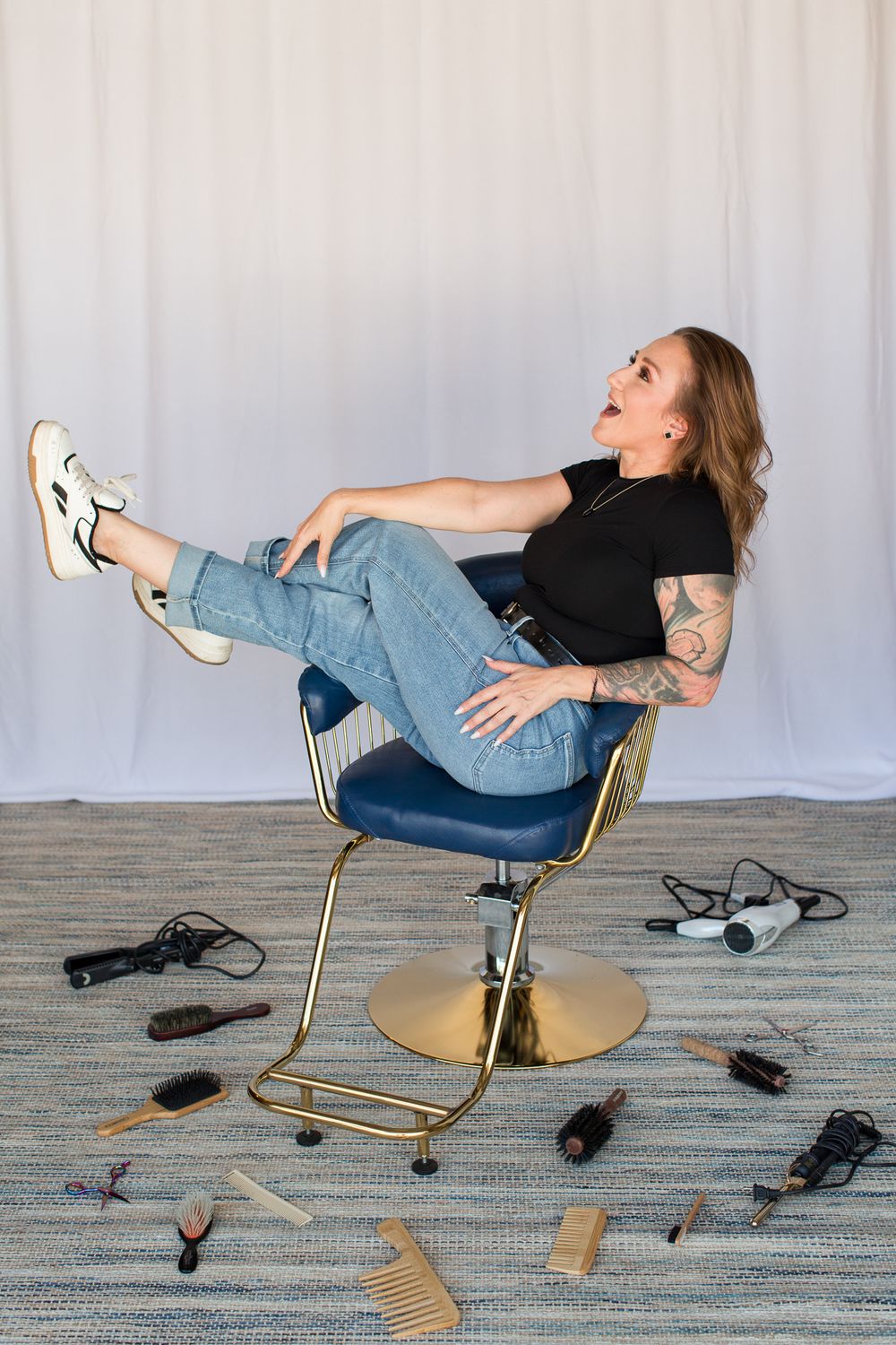 Playful pose in blue salon chair with various hair styling tools scattered on floor.