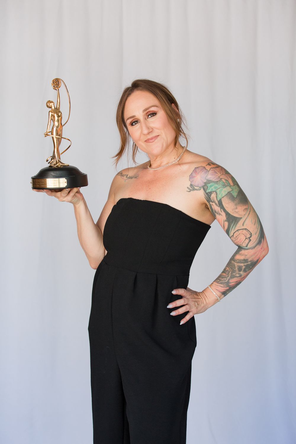 Figure in black dress poses with trophy and awards in studio setting.