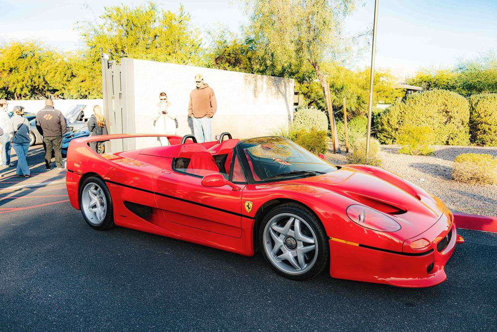 A bright red Ferrari F50 supercar parked on a sunny road with trees and foliage in the background.