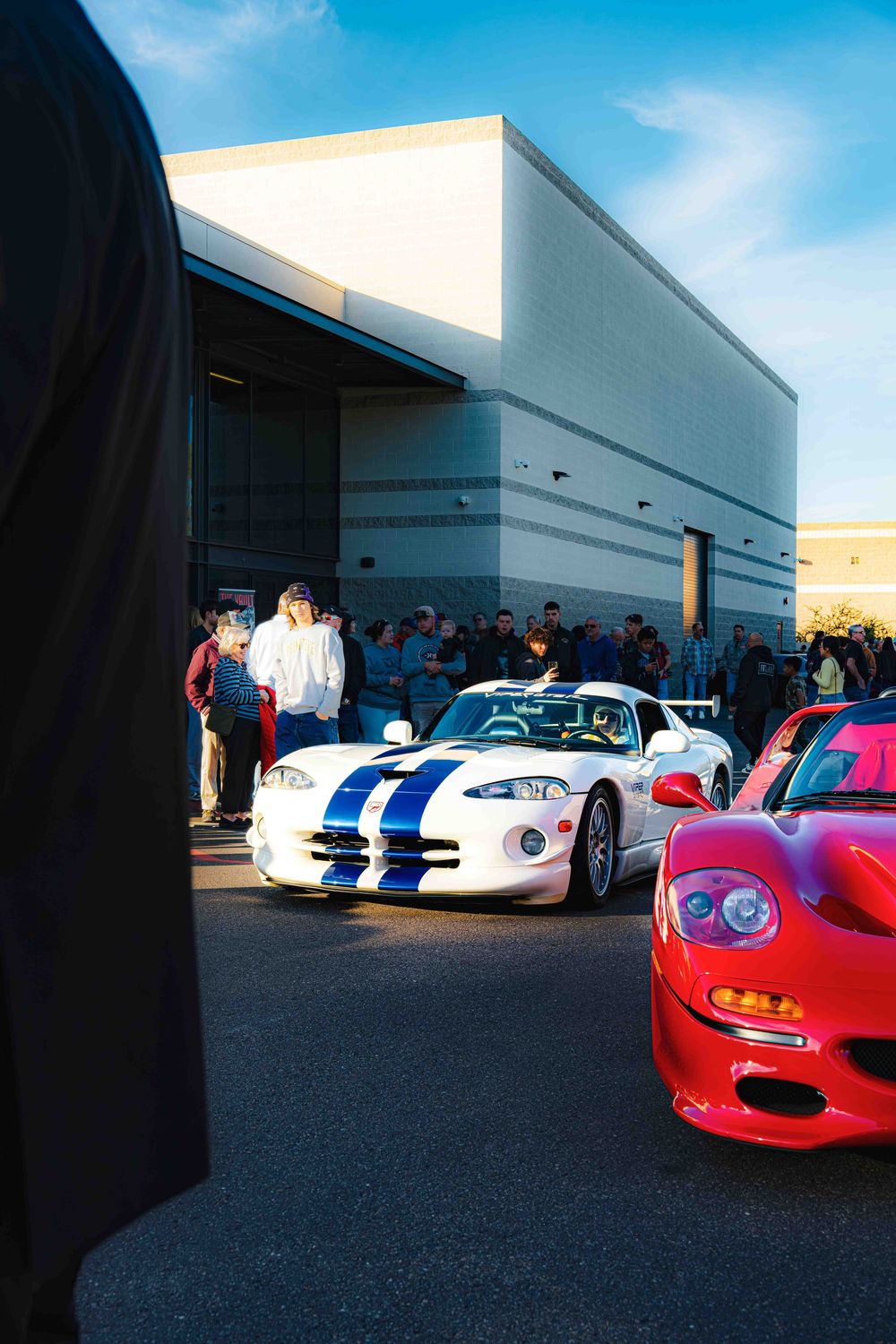 A white Dodge Viper with blue racing stripes alongside other sports cars at a car meet outside a building.