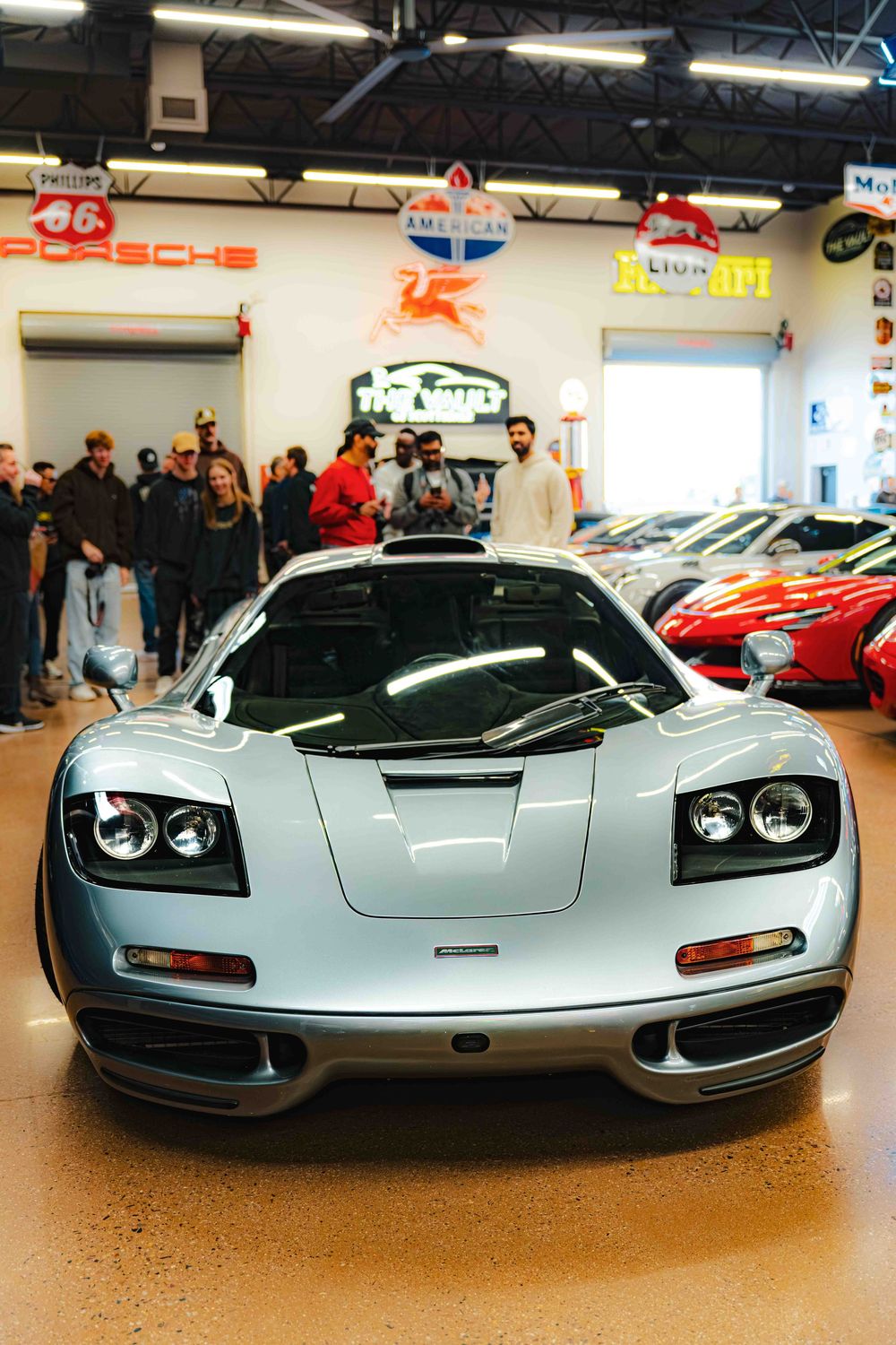 A silver McLaren F1 supercar on display in a showroom with spectators gathered around to view the iconic vehicle.