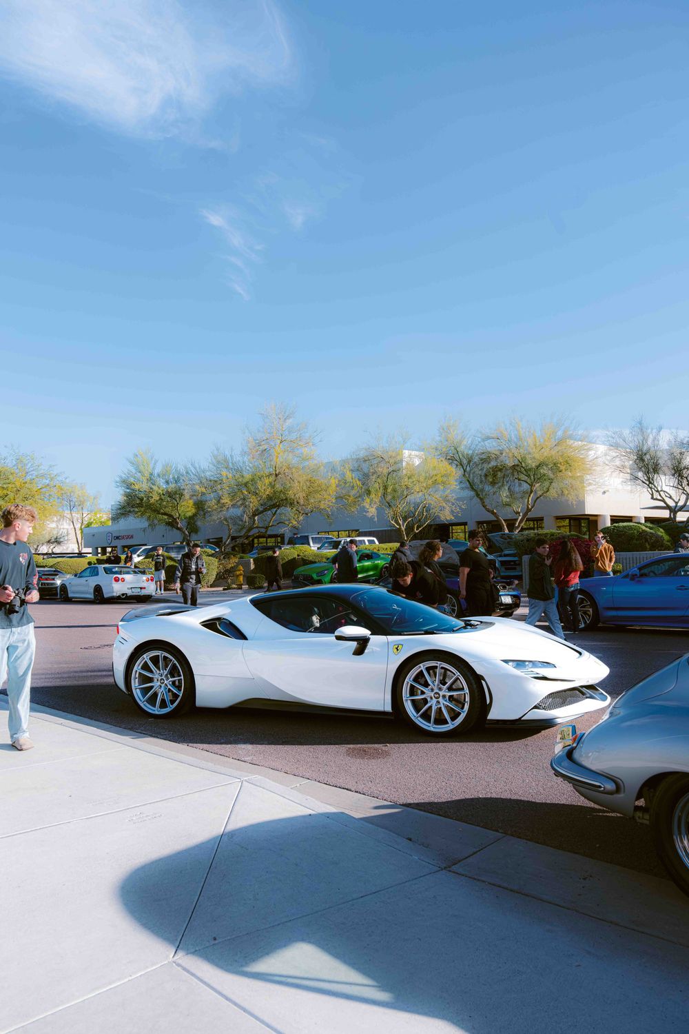 A white Ferrari SF90 Stradale supercar parked in a lot with other vehicles under a bright blue sky.