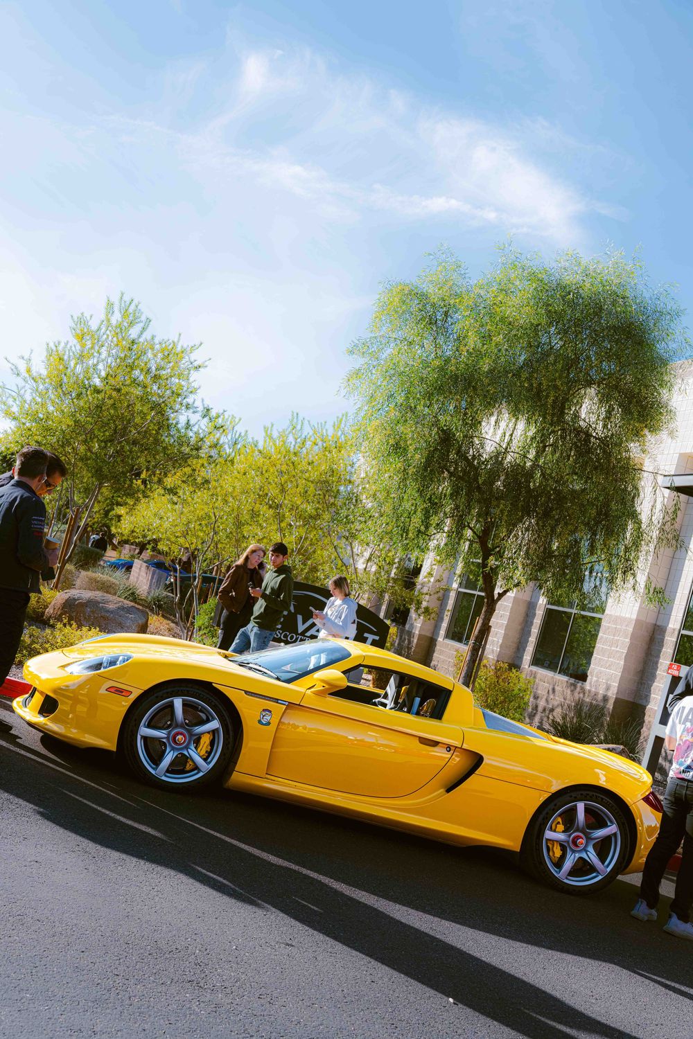 A bright yellow Porsche Carrera GT supercar parked on a street with trees and onlookers in the background.