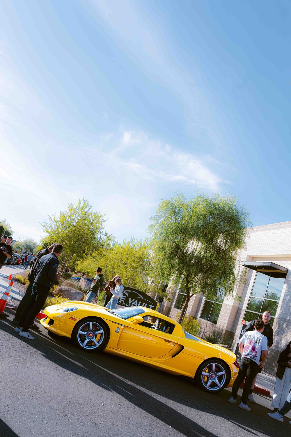 A bright yellow sports car parked outside a building draws attention from onlookers on a sunny day.