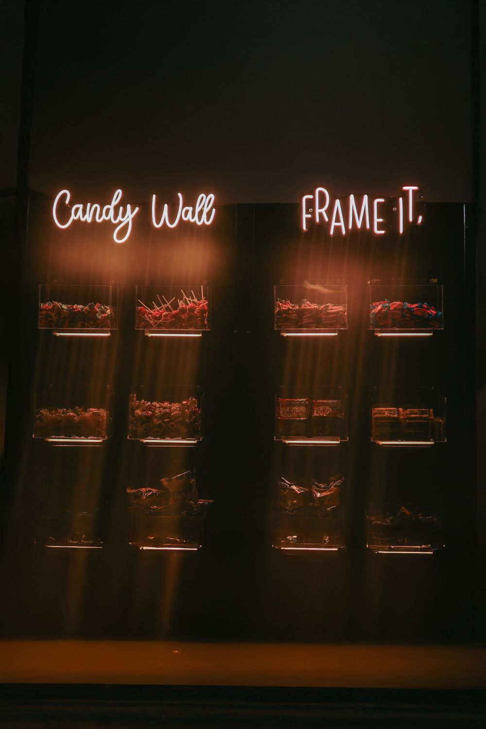 Illuminated neon signs reading 'Candy Wall' and 'Frame It' glow warmly against a dark wall display with shelving.