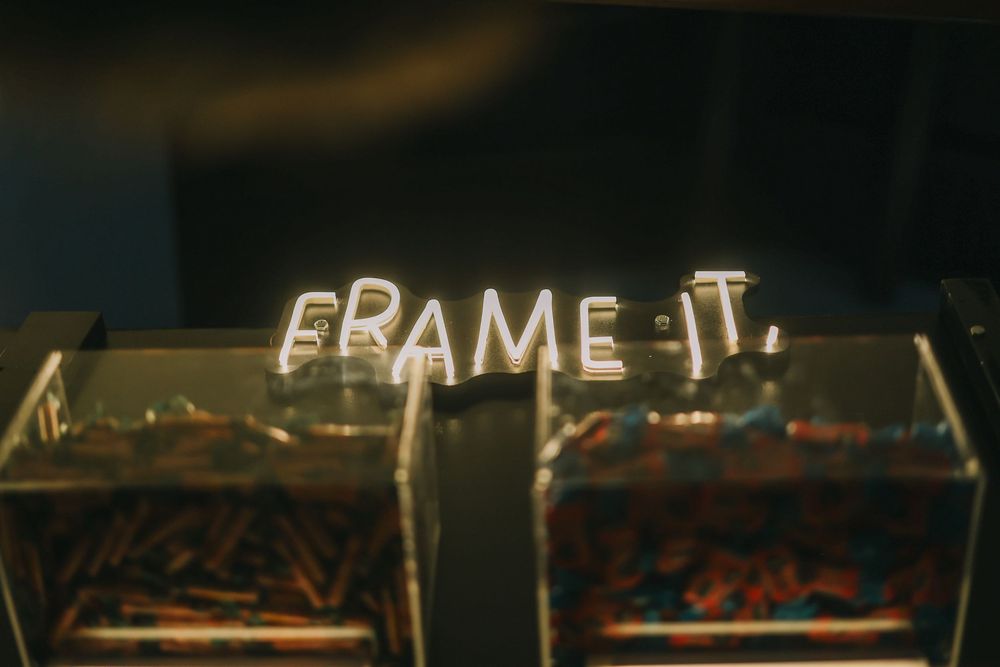 Illuminated neon text reading 'FRAME IT' glows against a dark background above what appears to be display cases.