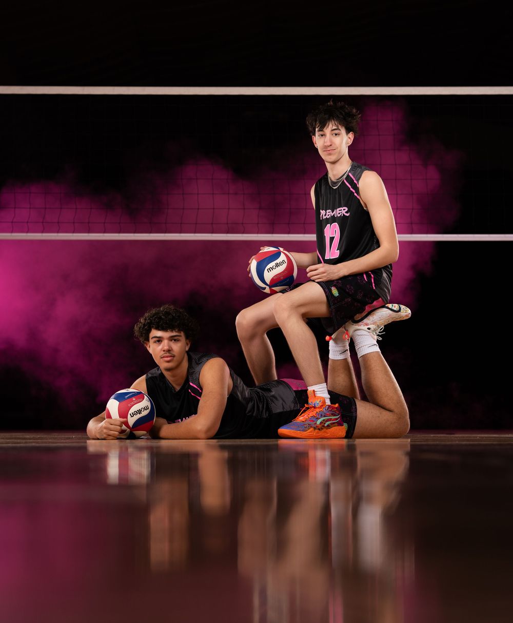 Two volleyball players pose with balls against a dark backdrop with pink smoke effects on a glossy court.