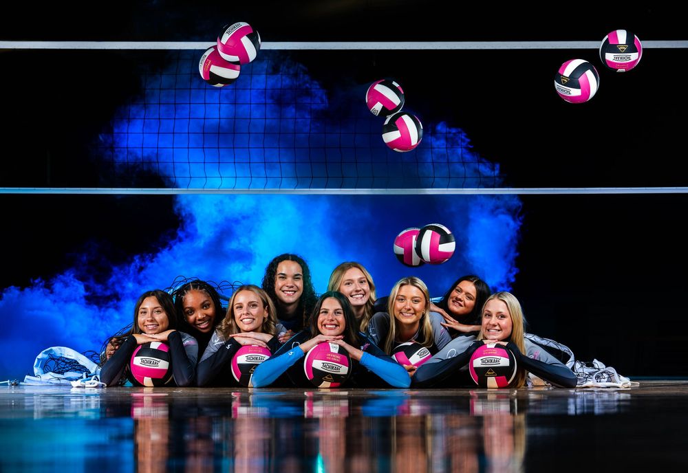 Volleyball players pose with pink volleyballs in a dramatic blue-lit sports arena setting.