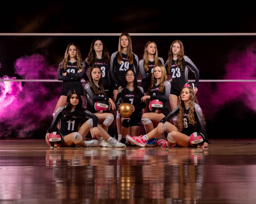 Volleyball team members sit on a wooden court floor in black uniforms with pink lighting effects in the background.