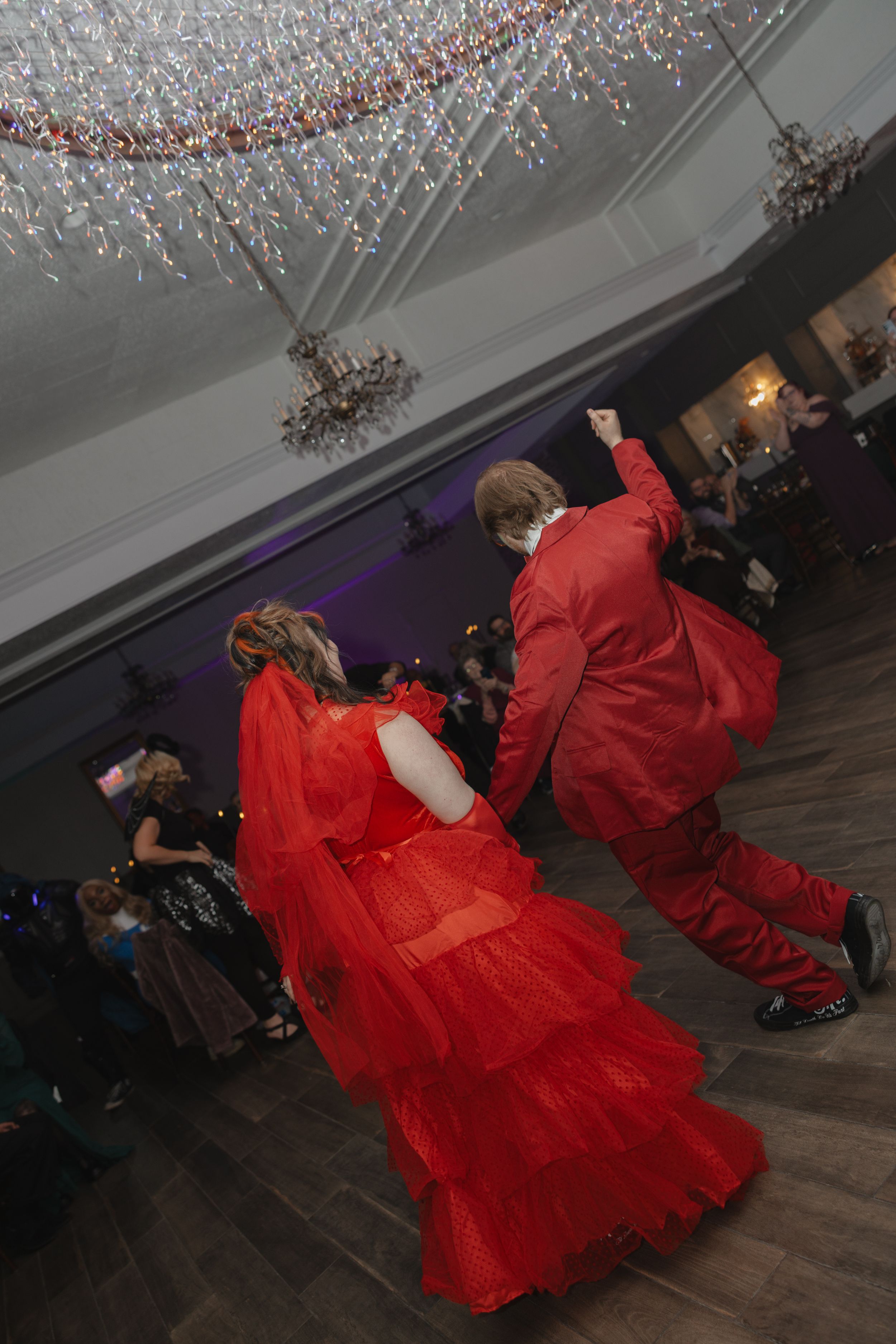A person in a flowing red dress spins and twirls on a wooden dance floor beneath sparkling decorative lights.