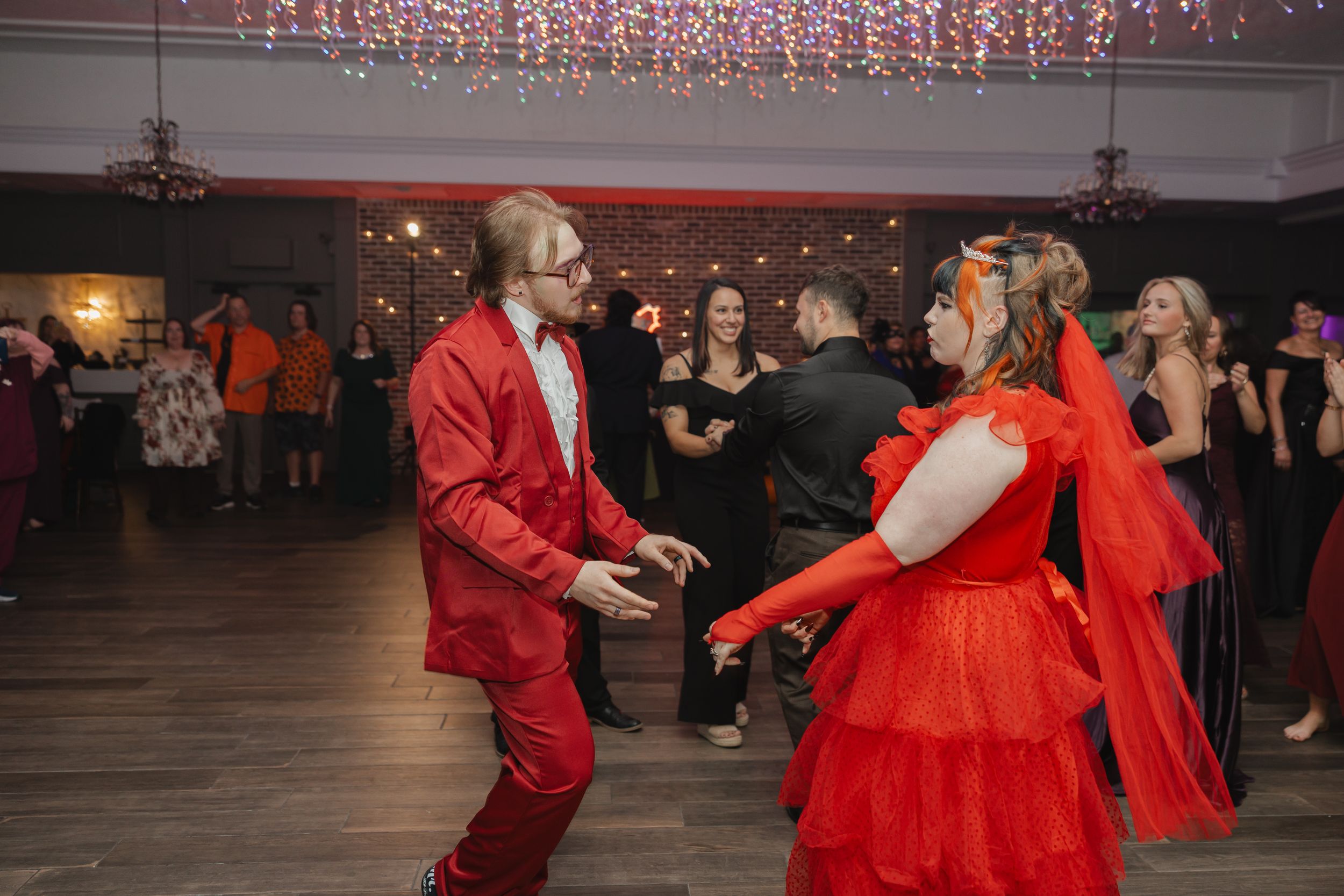 Dancers in vibrant red outfits perform together on a wooden dance floor under twinkling lights.