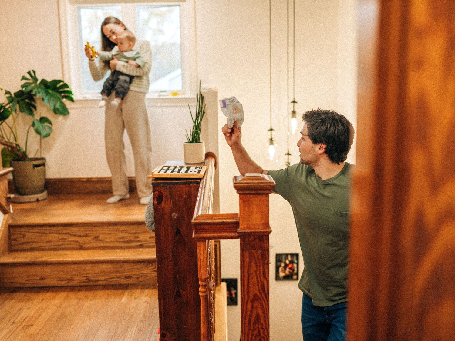 A person stands on wooden stairs in a sunlit room while holding a baby against their shoulder.