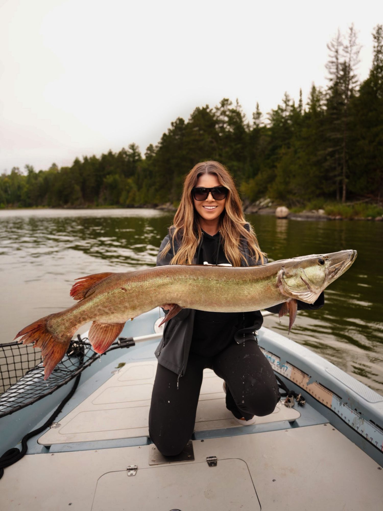 Impressive musky fish catch displayed on a boat with lake and forest in background.