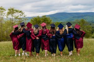 Group of graduates in maroon and navy gowns embrace while overlooking a scenic mountain meadow.