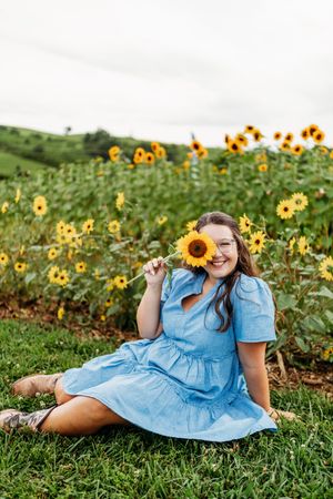 Smiling young woman in blue dress holding sunflower while sitting in sunflower field.
