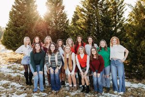 Group of teenage girls smiling together in front of snow-covered evergreen trees.