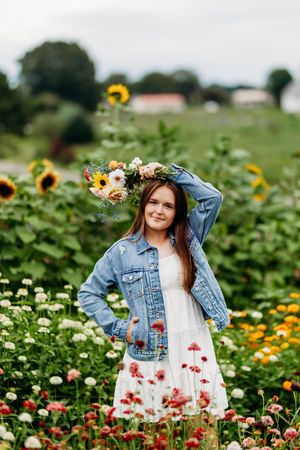 Young woman in white dress and denim jacket holding flower crown in a colorful flower field.