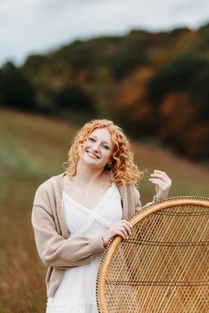 Smiling young woman with curly red hair poses beside a wicker peacock chair in an autumn field.