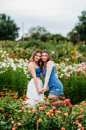 Two smiling young women hugging in a vibrant garden full of colorful blooms, wearing blue and white outfits.