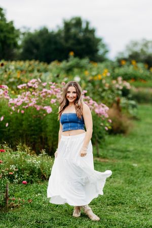 Smiling young woman in denim tube top and flowing white skirt twirling in a flower garden.