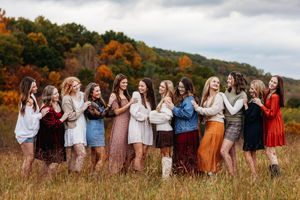 Group of eleven young women in colorful fall outfits pose together in a scenic autumn meadow.