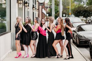 Group of seven women posing on a sidewalk wearing black dresses with pink accents for a bachelorette party.