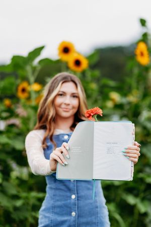 Young woman holding open journal with orange flower inside in a sunflower field.