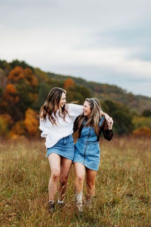 Two laughing young women in denim skirts walking through golden autumn meadow.