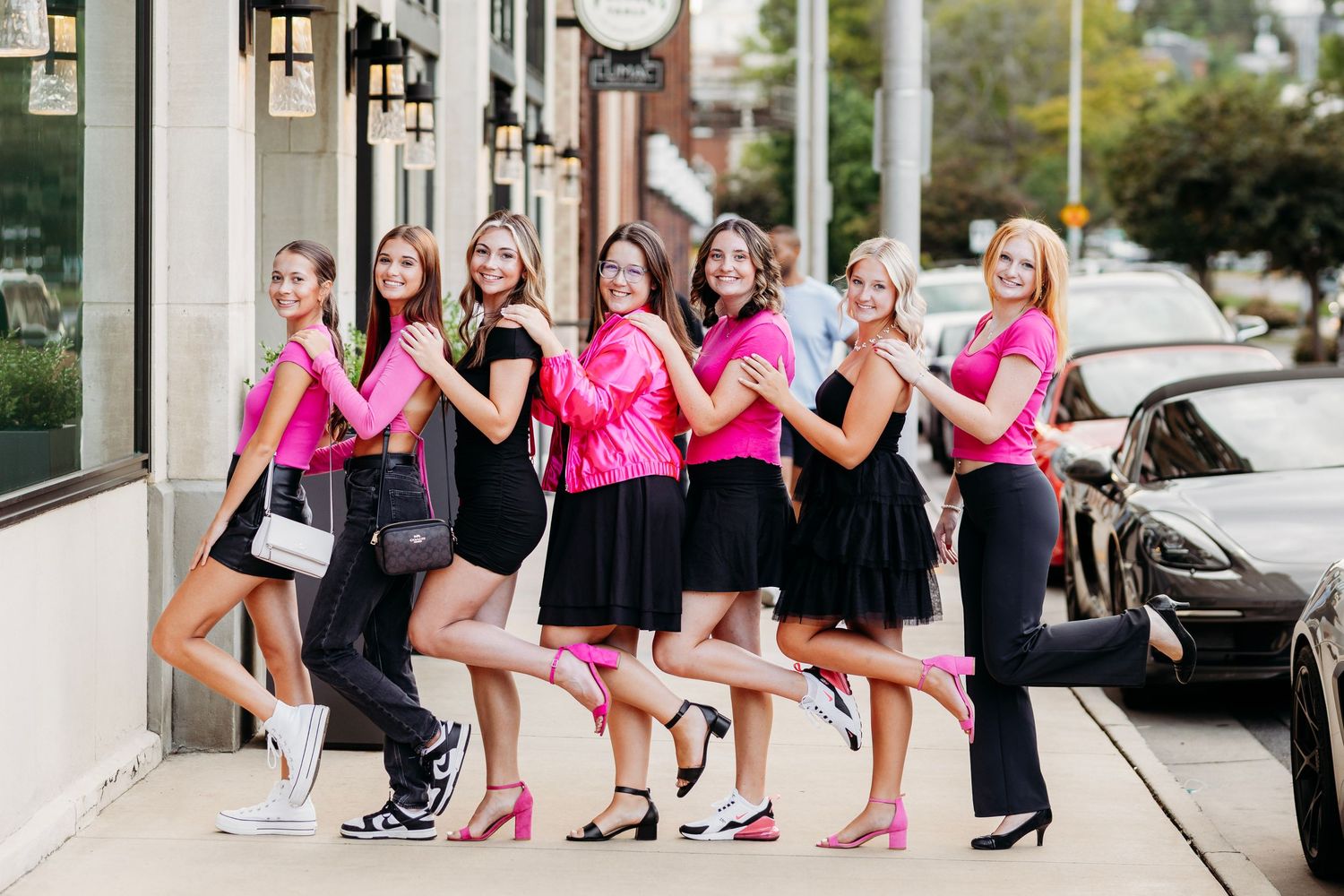 Seven women in matching pink and black outfits posing playfully on a city sidewalk, kicking legs up.