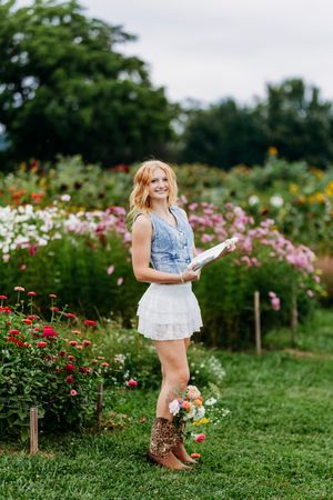 Smiling blonde woman in denim vest and white skirt holding book in flower garden.