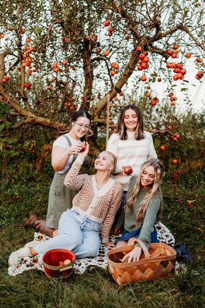 Four women enjoying a picnic under apple trees in an orchard, holding fresh red apples on a blanket.
