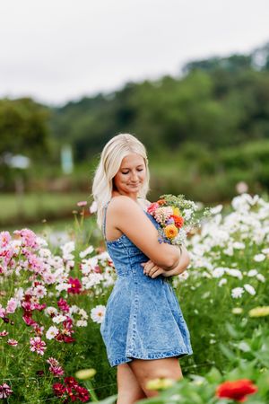 Blonde woman in denim dress holding colorful bouquet in vibrant flower field.