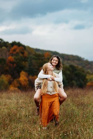 Two smiling young women pose playfully in a golden autumn meadow, one carrying the other on her back.