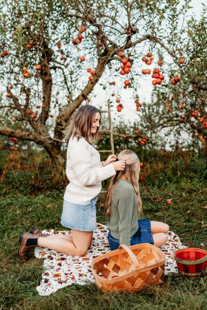 Two young women on a blanket in an apple orchard, one braiding the others long blonde hair.
