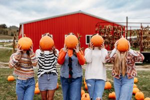 Five people holding large orange pumpkins in front of their faces near a red barn at a pumpkin patch.