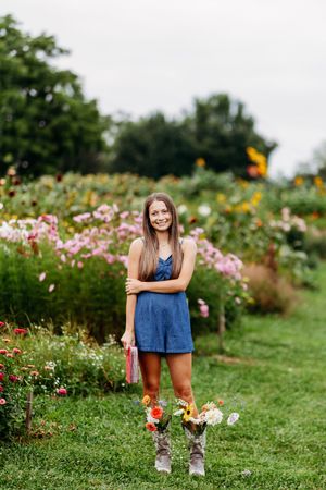 Brunette woman in blue romper smiling in colorful flower garden holding bouquet.
