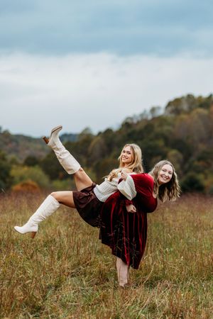 Two young women laugh in an autumn field, one giving the other a playful piggyback ride in fall fashion.