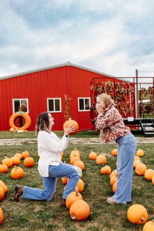 Woman kneeling with a pumpkin in a playful proposal pose at a fall pumpkin patch near a red barn.